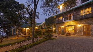 A two-story building with a stone facade, a balcony with a black railing, and a gravel driveway leading to a seating area with a table and chairs. - Seclude Nahan Bantony Cottage (Paonta Sahib)
