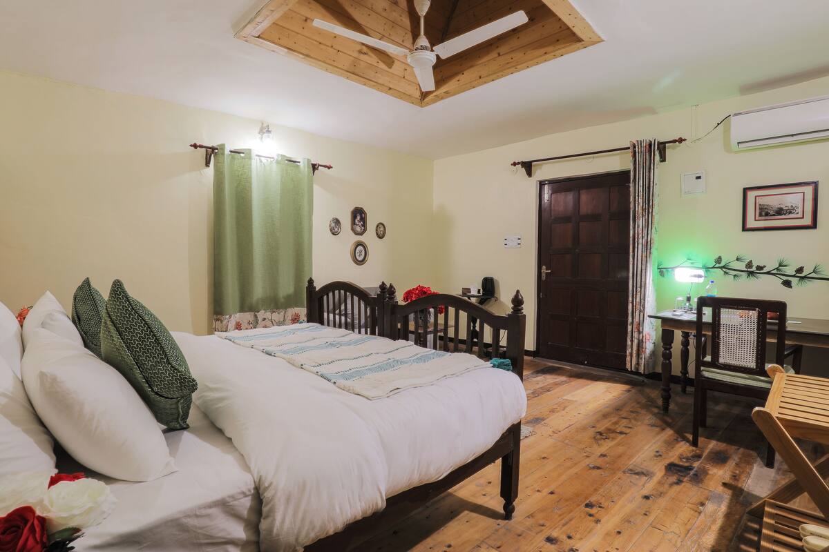 A room with a wooden bed, a desk, and a chair. There is a ceiling fan, a door, and a wall-mounted air conditioner.