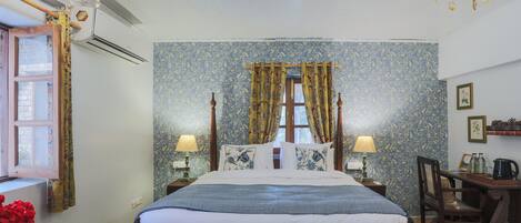 Un dormitorio ordenado con una cama, dos mesitas de noche con lámparas, una silla de madera, un escritorio con un hervidor y una ventana con cortinas.