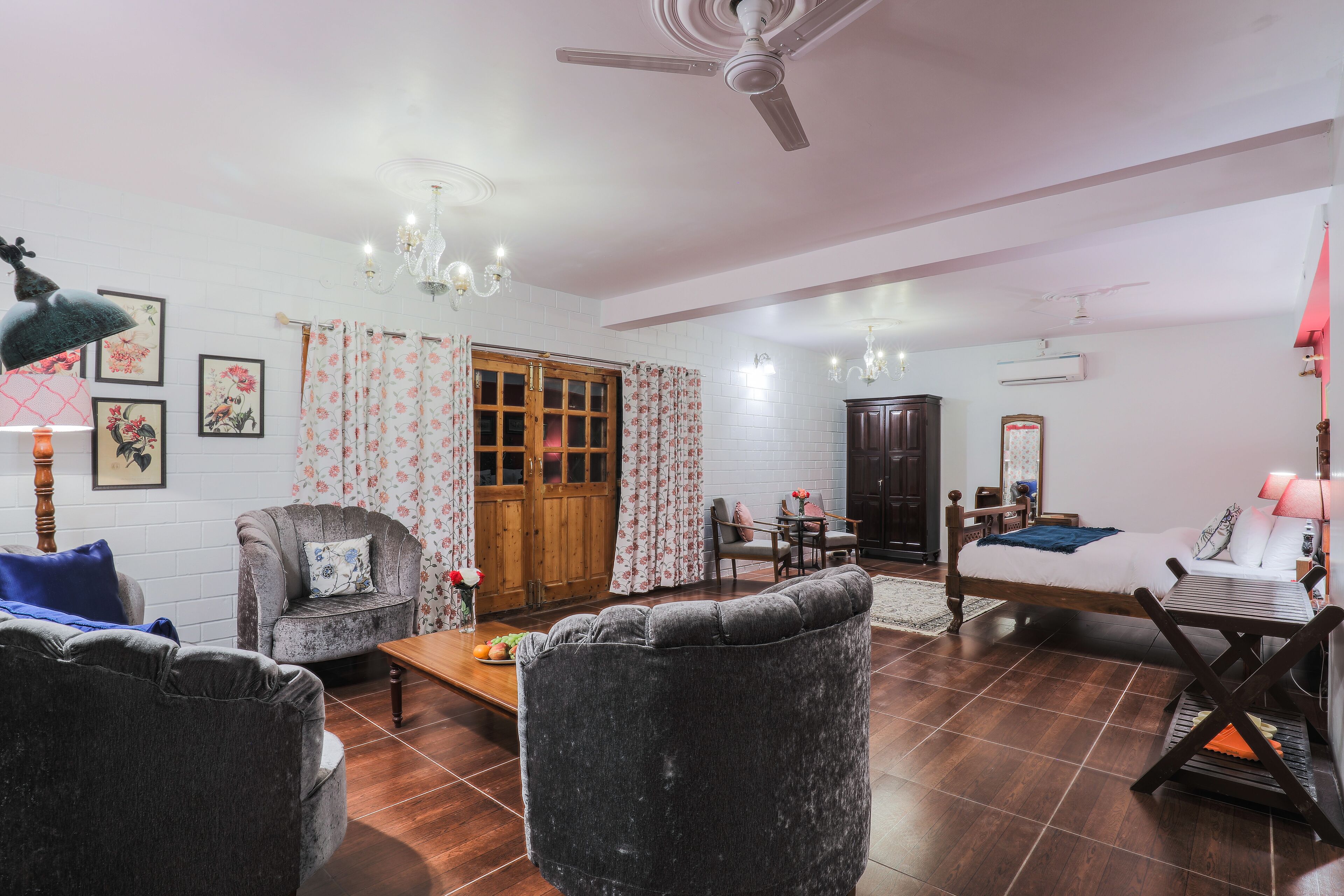Un salón espacioso con sofá, sillón y una mesa de centro de madera. Se observa una puerta de madera con cortinas florales, un candelabro y un ventilador de techo.