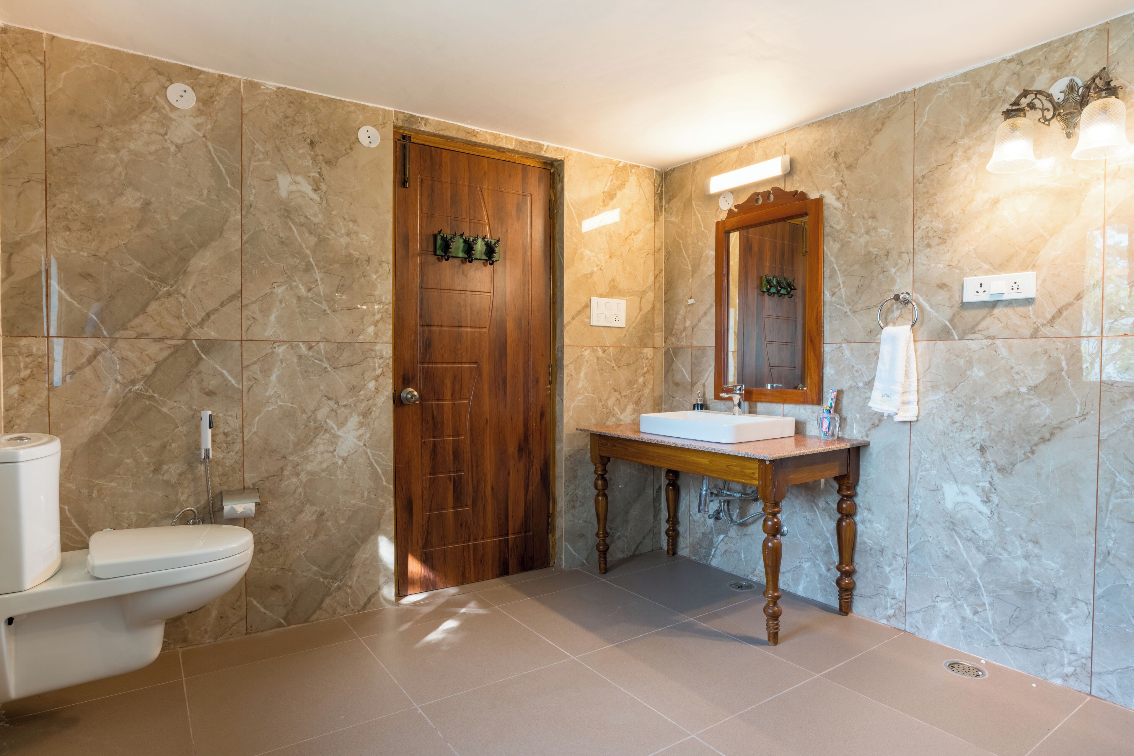 Un baño con puerta de madera, inodoro blanco, pared de mármol, tocador de madera con espejo y lavabo.