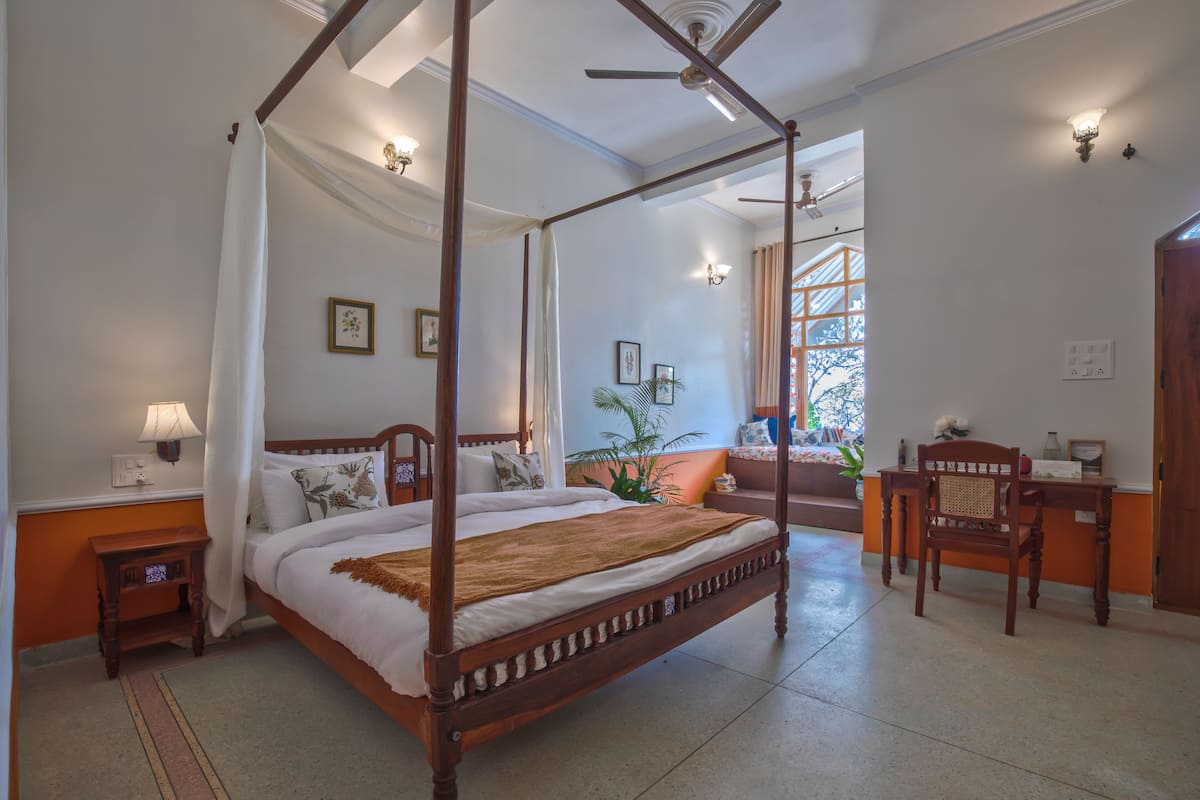 A four-poster bed with a canopy, a wooden nightstand, a desk with a chair, and a view of a balcony with a plant.