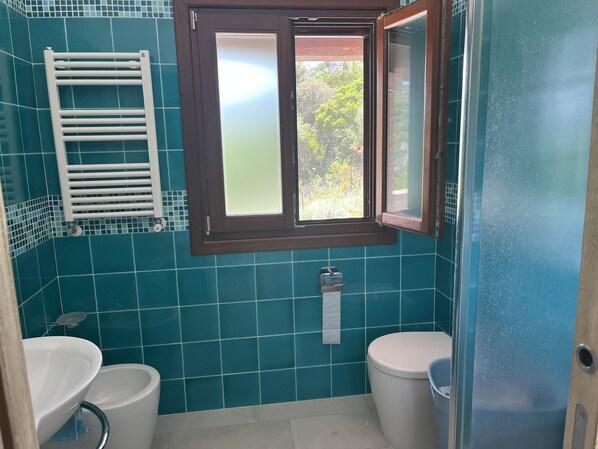 A bathroom with teal tiles, a white sink, a toilet, a window with a view of greenery, and a towel rack.