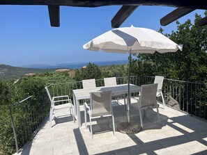 A terrace with a table and chairs, an umbrella, and a view of the sea and hills.