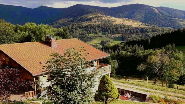 Uma casa com telhado inclinado, rodeada por vegetação exuberante e montanhas ao fundo.