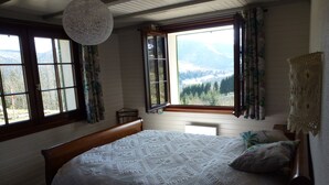Quarto com cama grande, cabeceira de madeira e vista para montanhas através de janelas abertas.
