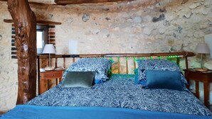 Un dormitorio rústico con pared de piedra, vigas de madera, colcha estampada y mesita de noche con lámpara.