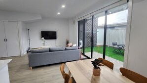 Een moderne woonkamer met een grijze bank, een flatscreen televisie en een eethoek met een houten tafel en stoelen.