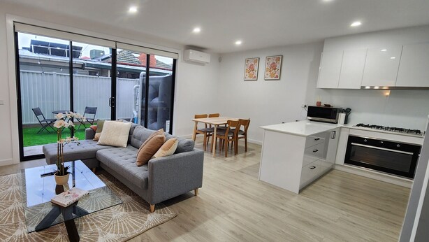 Een modern appartement met een grijze bank, een glazen salontafel, een eethoek met een tafel en stoelen, en een keuken met witte kasten en een fornuis.