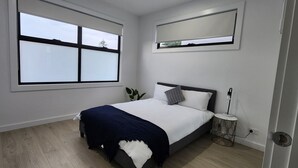 Een slaapkamer met een groot raam, een bed met een donkere deken, een nachtkastje met een lamp en een wandplank.