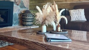 Un intérieur rustique avec une cheminée en pierre, des poutres en bois et un coin salon confortable agrémenté d’un coussin sur lequel est inscrit : « NOTRE PETIT COIN DE BONHEUR ».