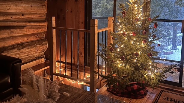 L’intérieur d’un chalet chaleureux, avec un mur en rondins de bois, un banc en bois, un sapin de Noël illuminé et une fenêtre donnant sur un paysage enneigé.
