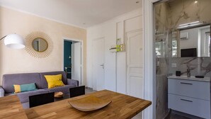 A modern living room with a wooden dining table, a gray sofa with yellow cushions, a round mirror, and a kitchen area with marble countertops and a sink. - 2 Pieces Balcon et Climatisation 32m2 (Cannes)