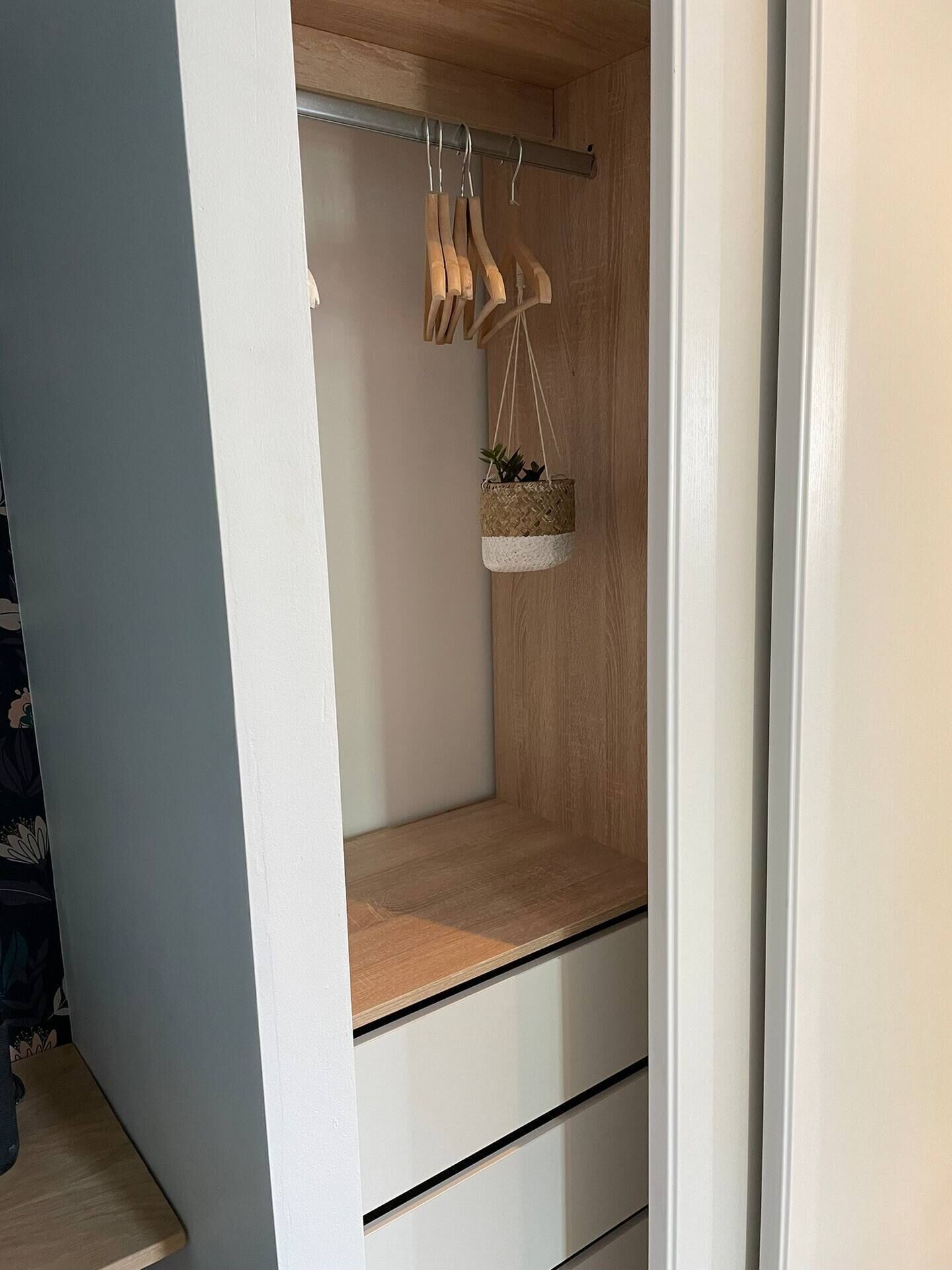 Un vestidor con estantes de madera, una barra para colgar con perchas de madera y una pequeña cesta tejida con una planta.