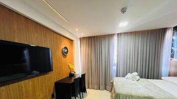 A hotel room with a flat-screen TV mounted on the wall, a desk with a chair, a bed with pillows, and a large window with curtains.