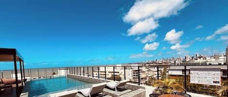 Piscina no terraço com espreguiçadeiras e uma mesa, com vista para a paisagem urbana.