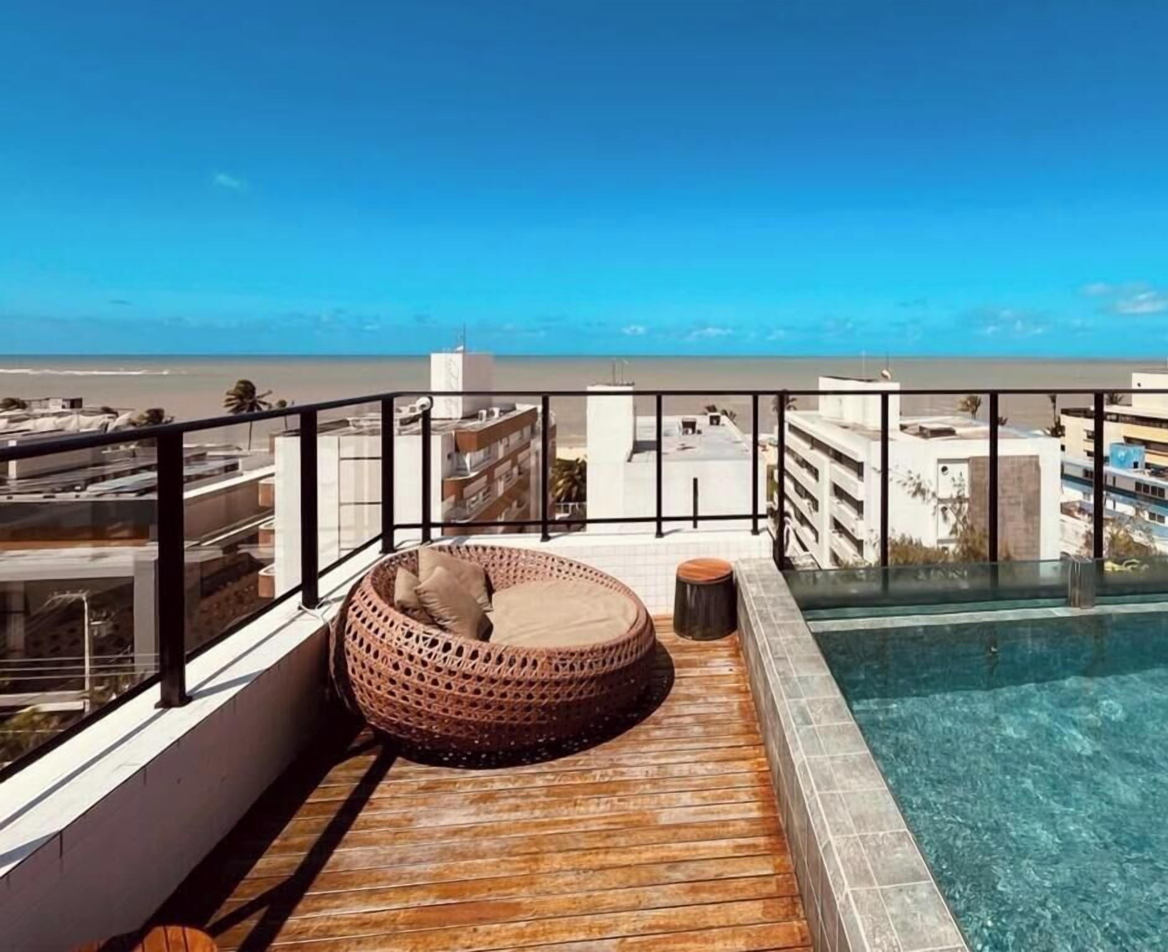 A rooftop pool area with a clear view of the beach, a wooden deck, and a wicker lounge chair.