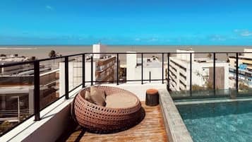 A rooftop pool area with a clear view of the beach, a wooden deck, and a wicker lounge chair.