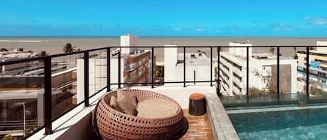 A rooftop pool area with a clear view of the beach, a wooden deck, and a wicker lounge chair.