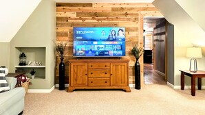 A living room with a flat-screen TV on a wooden cabinet, a small shelf with a 'Relax' sign, a vase with branches, and a lamp on a table.