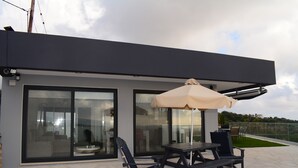 Un patio exterior moderno con una mesa, sillas, un paraguas grande y un edificio con cerramientos de vidrio.