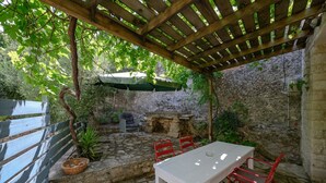 A patio area with a stone floor, a table with chairs, a pergola with a green umbrella, and a stone wall in the background. - Rogalida,serene villa,pool,Near taverns (Lampini)