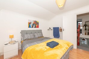 A bedroom with a bed, a bedside table, a lamp, a painting, and a towel rack. - Spacious 4BR London Home Near Train, ExCeL & O2 (London)