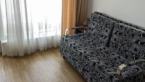 A room with a sofa, wooden flooring, a window with sheer curtains, and a wall-mounted air conditioning unit. - Beautiful 1-bed Studio in Sunny Beach Bulgaria (Nessebar)