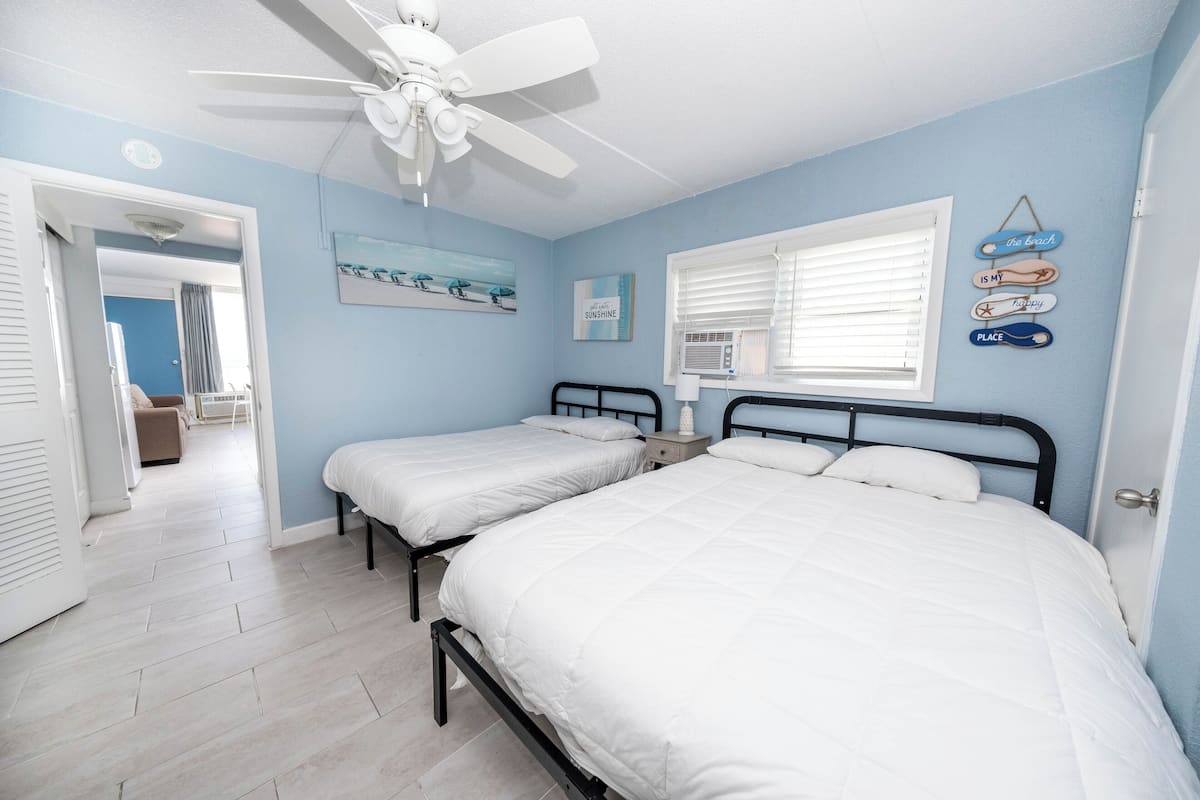 Une chambre avec deux lits, un ventilateur de plafond, une fenêtre avec des stores et des décorations murales.