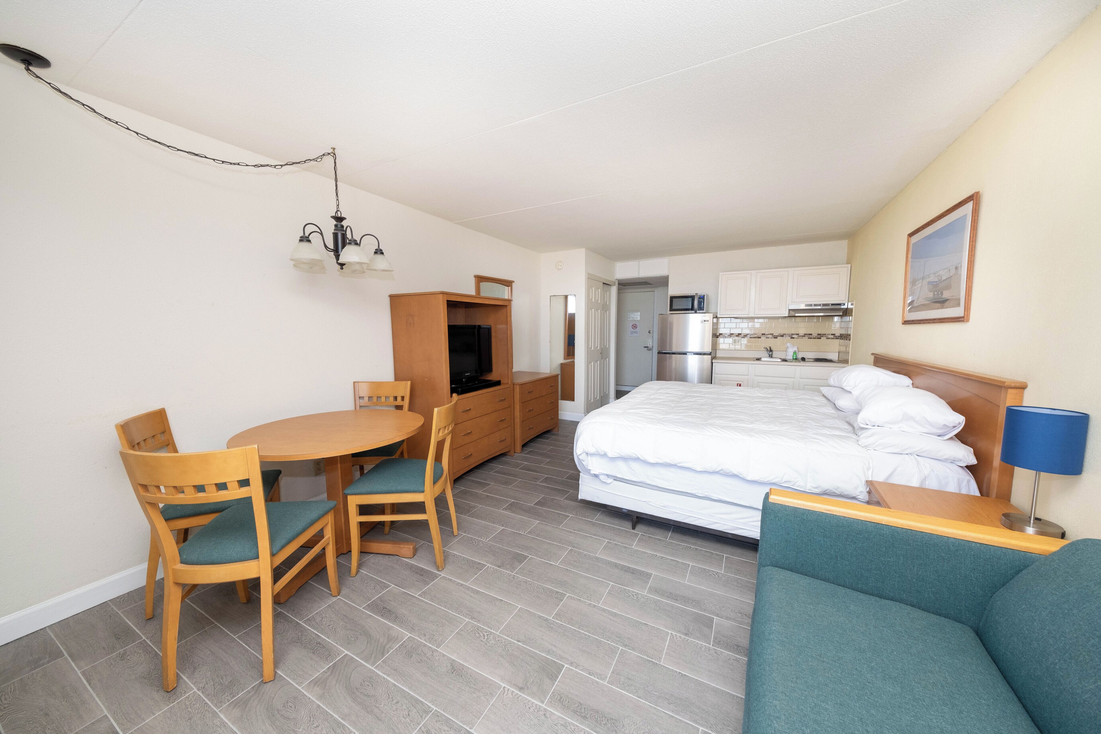 A compact hotel room with a bed, a small dining table with chairs, a kitchenette, and a TV.