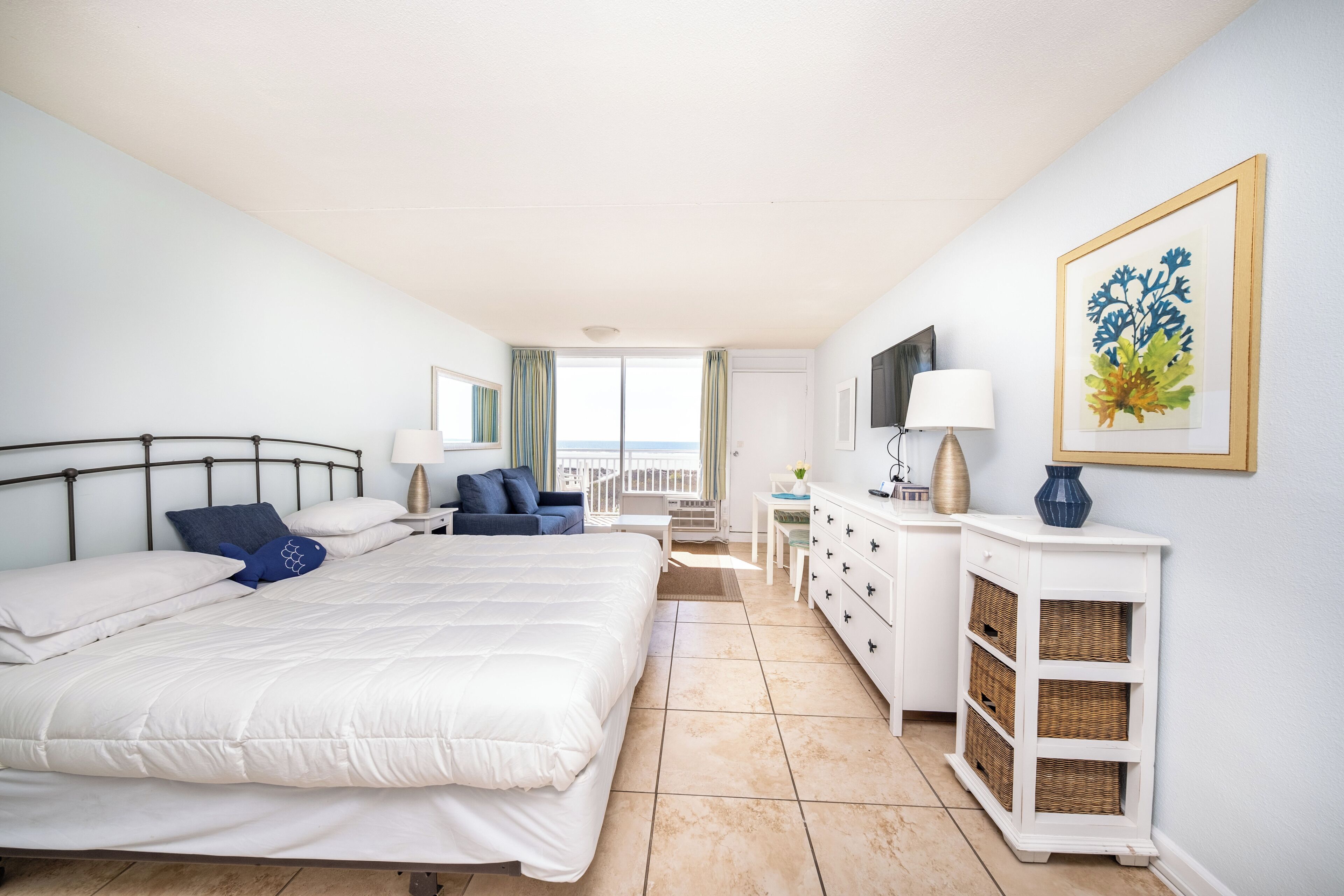 A hotel room with a large bed, a desk, a chair, a TV, a painting, and a view of the sea.