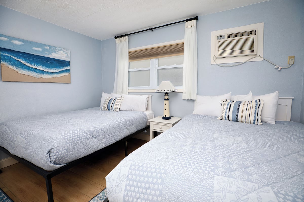 Une chambre avec deux lits, une table de chevet, une lampe, un tableau représentant une plage et une fenêtre avec des rideaux.