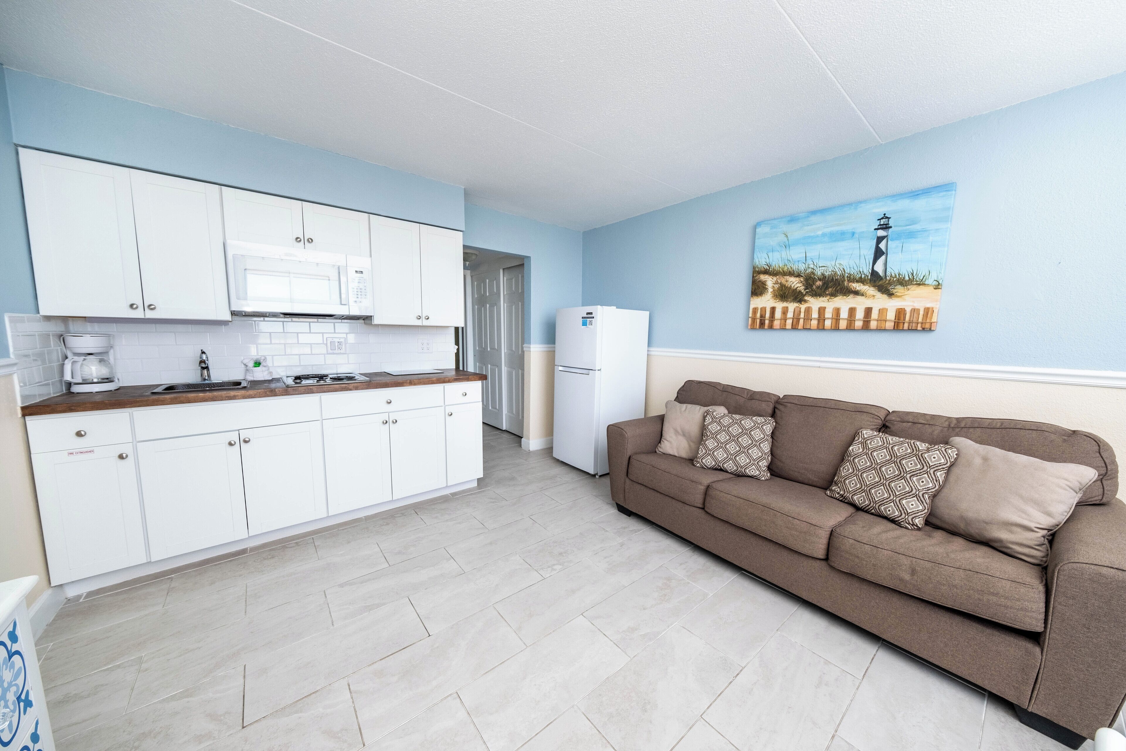Une cuisine moderne avec des meubles blancs et un four à micro-ondes, un canapé beige agrémenté de coussins à motifs et un tableau représentant un phare accroché au mur.