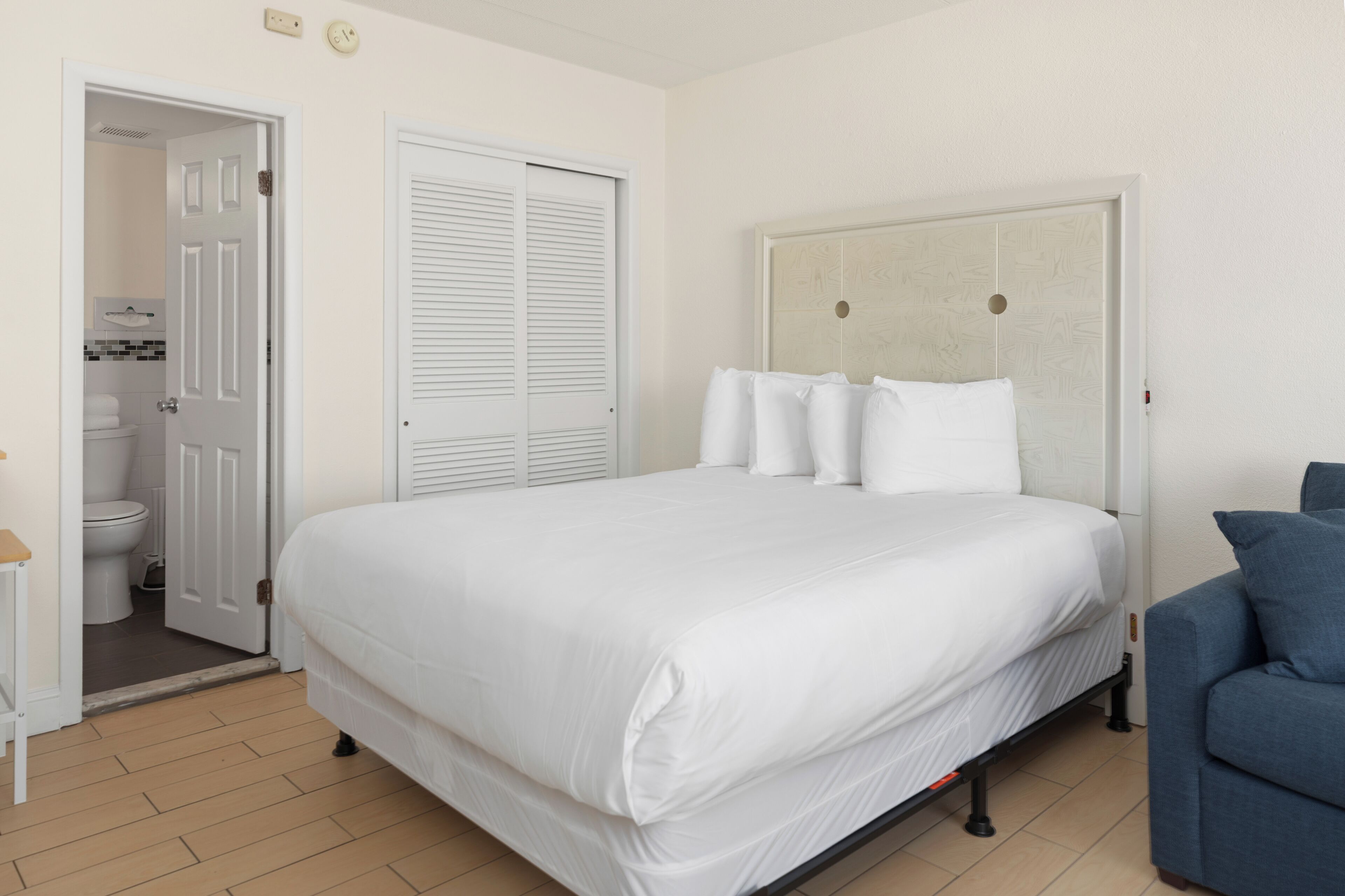 A bedroom with a bed, a blue chair, a white wardrobe, and a bathroom visible through an open door.