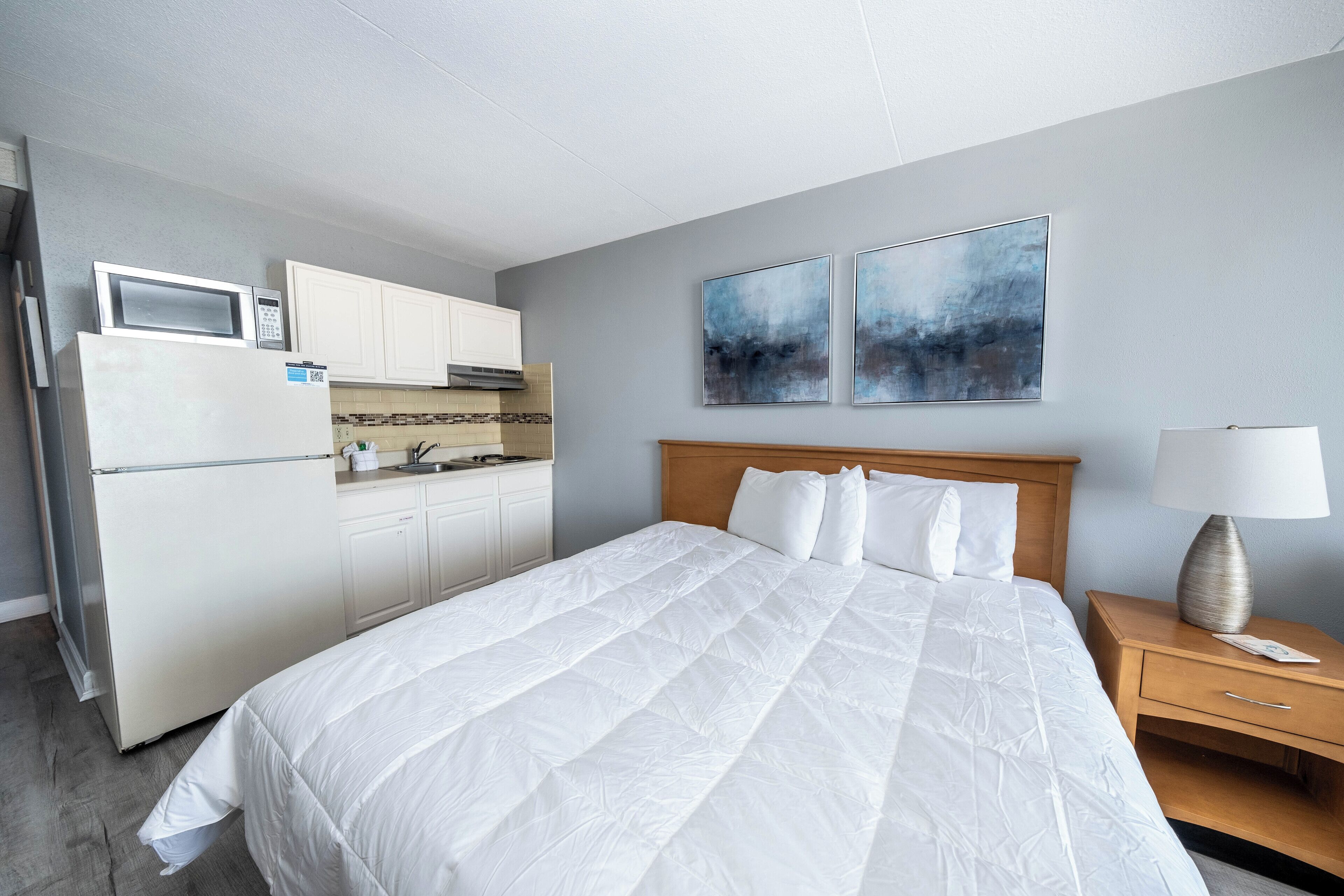Una habitación de hotel con cama, mesita de noche, lámpara, microondas y kitchenette.