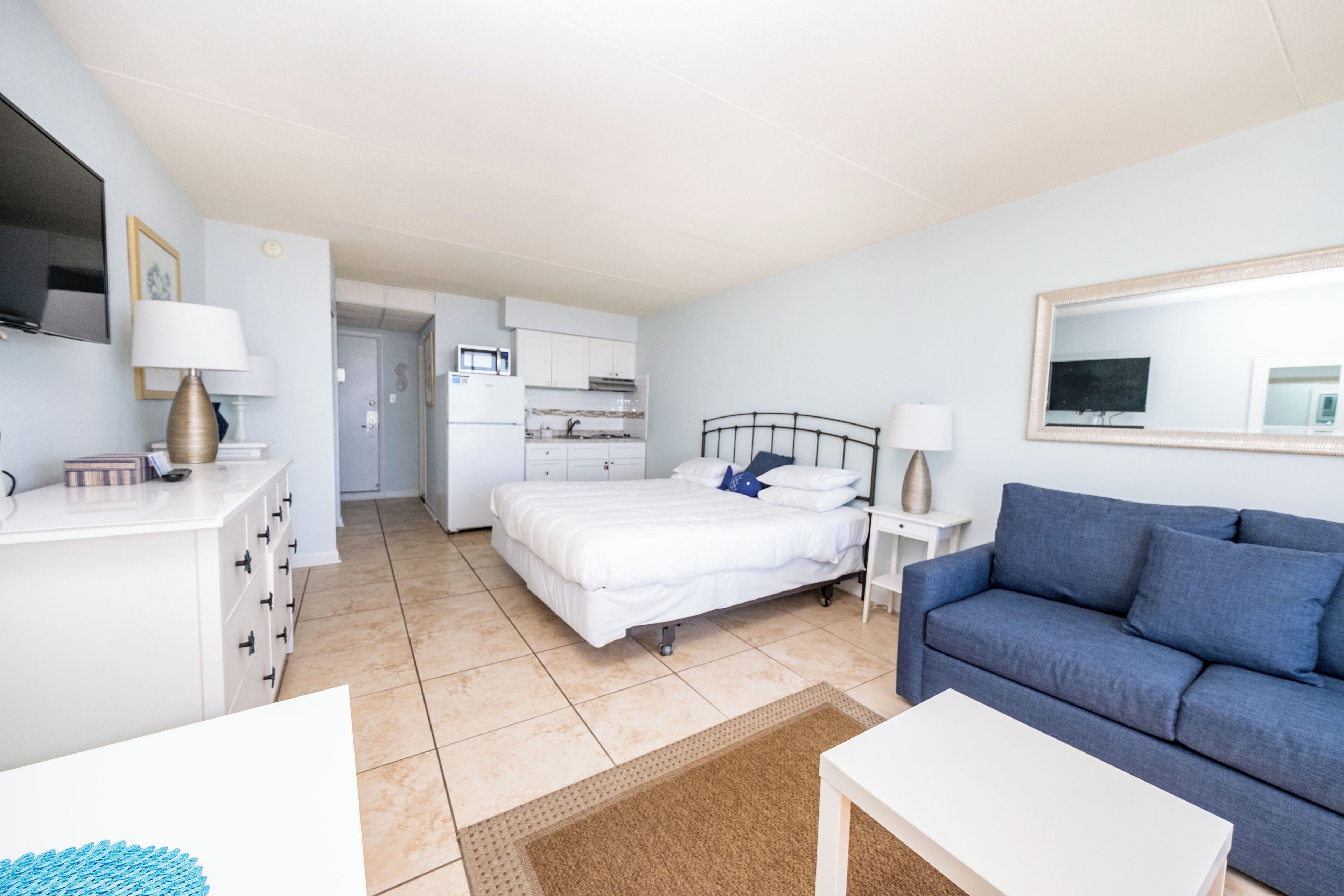 Un dormitorio con cama, sofá azul, mesita de noche blanca, televisión y espacio de cocina.
