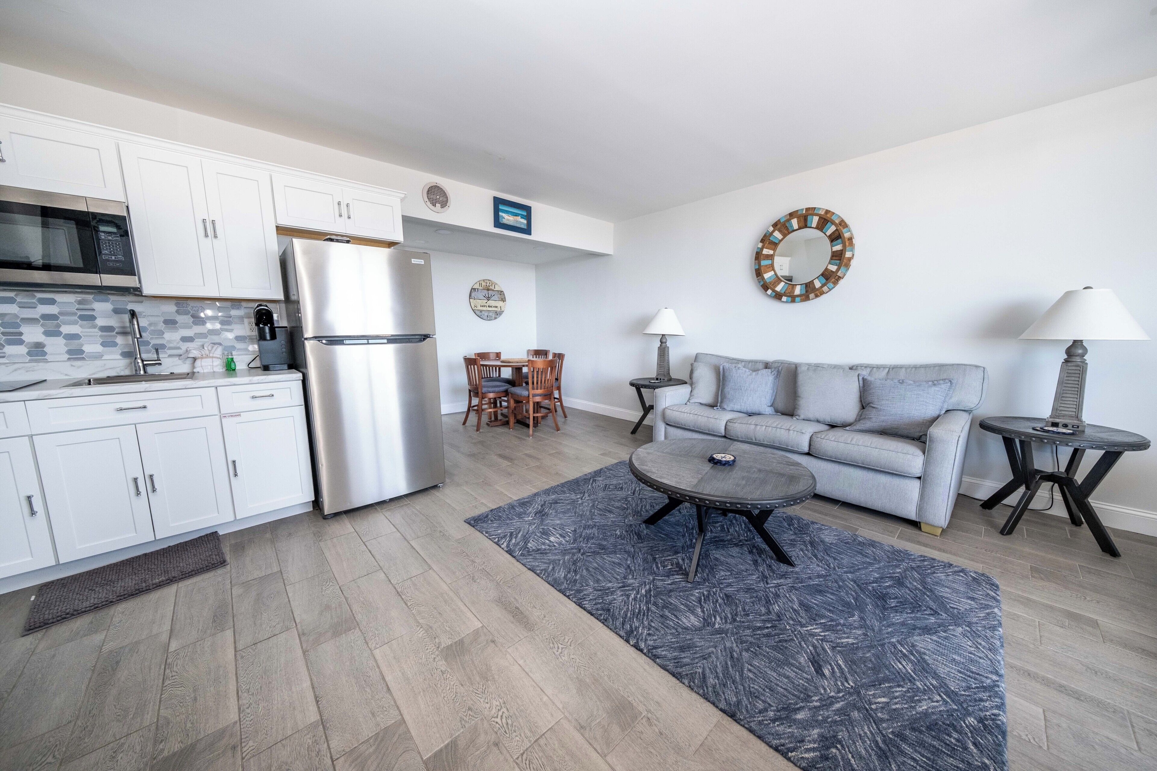 A modern kitchen with white cabinets, a stainless steel refrigerator, and a microwave. There is a dining area with a table and chairs, and a gray sofa with a matching throw rug.