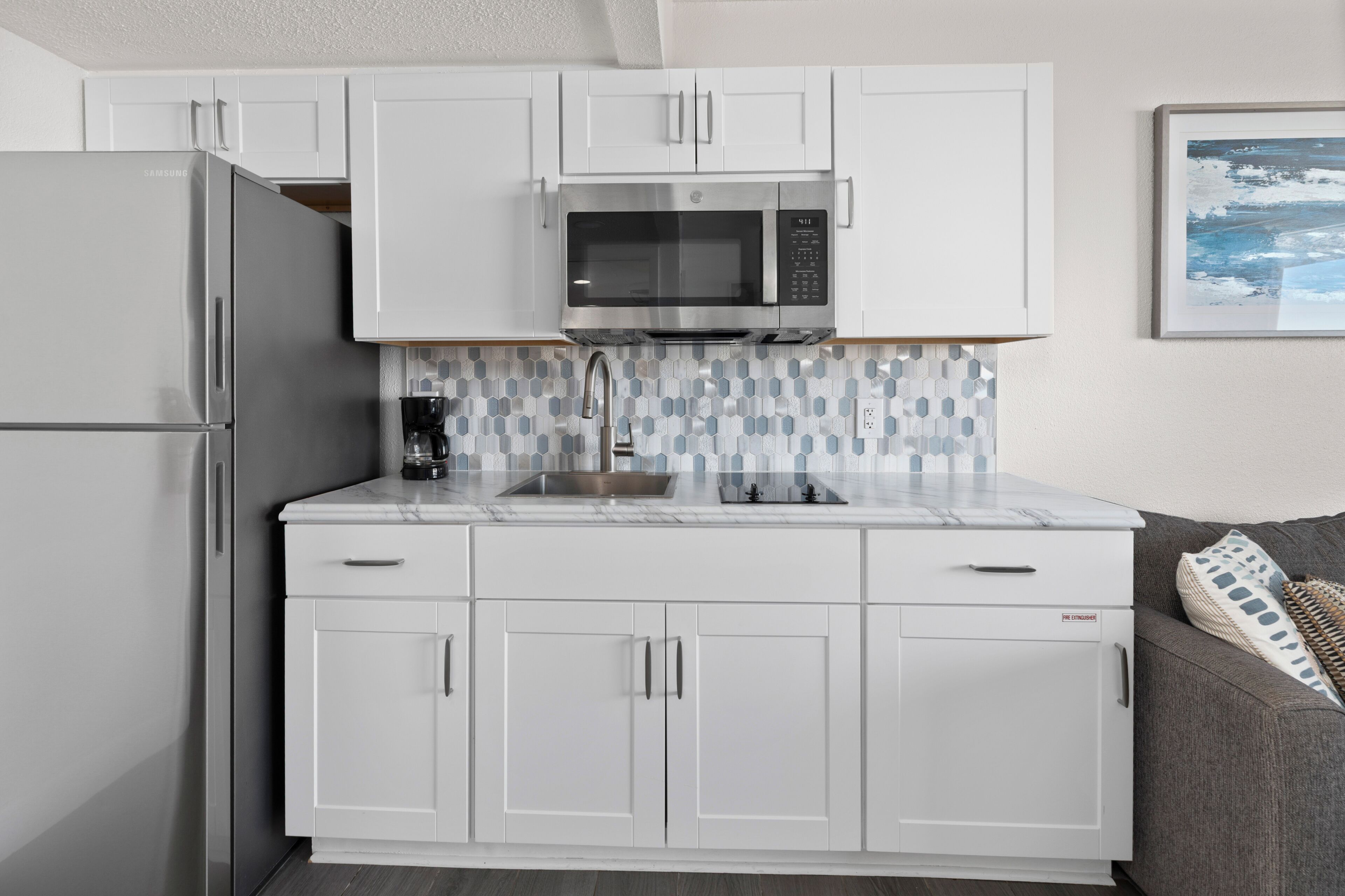 Una cocina compacta con armarios blancos, un refrigerador de acero inoxidable, un microondas y una cafetera.