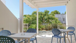 An outdoor patio with white tables and blue chairs, surrounded by greenery and a view of a building.