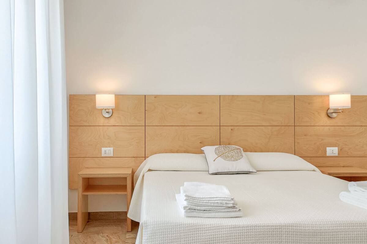 A hotel room with a wooden headboard, two bedside tables, a bed with white bedding, and two wall-mounted lamps.