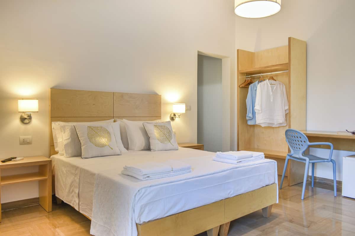 A neatly made bed with white linens and decorative pillows, a wooden nightstand with a lamp, a desk with a chair, and a wooden wardrobe with hanging clothes.