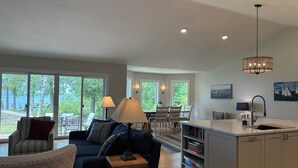 A spacious living room with a large window, a dining area, and a kitchen with a white countertop and cabinets. - Enjoy nightly sunsets at this Beautiful Door County Waterfront Home (Sturgeon Bay)