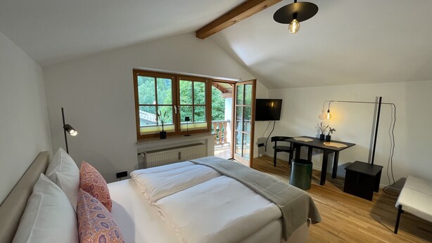 Un dormitorio con cama, escritorio, silla, televisor y ventana con vista a follaje.