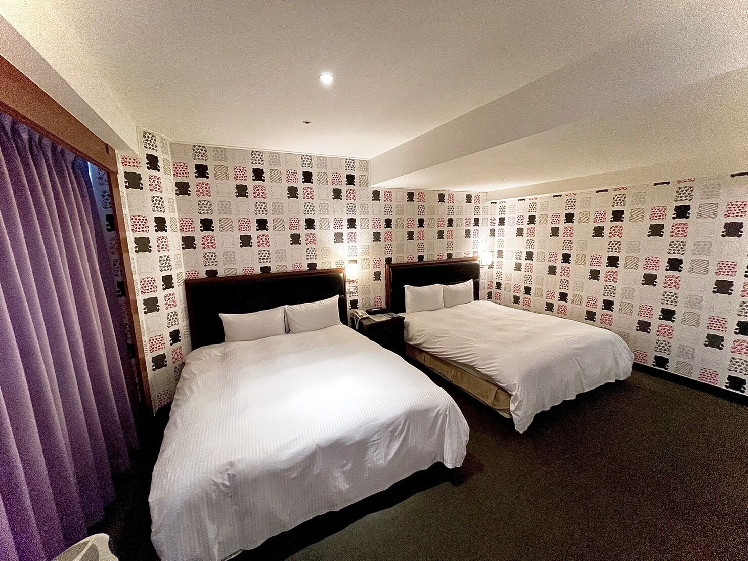 A hotel room with two beds, a wooden headboard, a nightstand, and a patterned wallpaper.