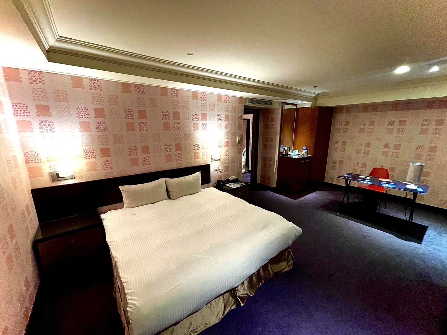 Habitación de hotel con una cama grande, un escritorio y una silla. Hay un espejo y una lámpara sobre el escritorio.