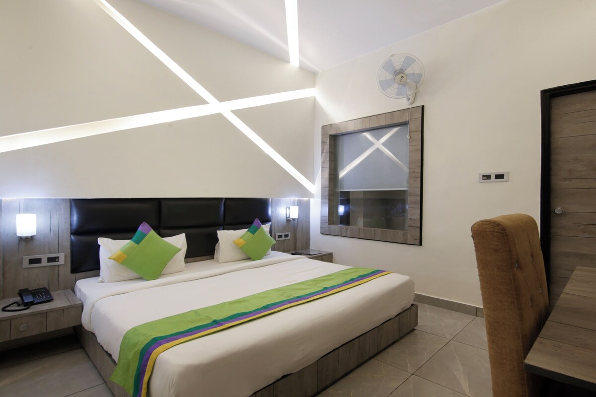 A modern hotel room with a large bed, a wooden headboard, a bedside table with a phone, a wall-mounted fan, and a wooden door.