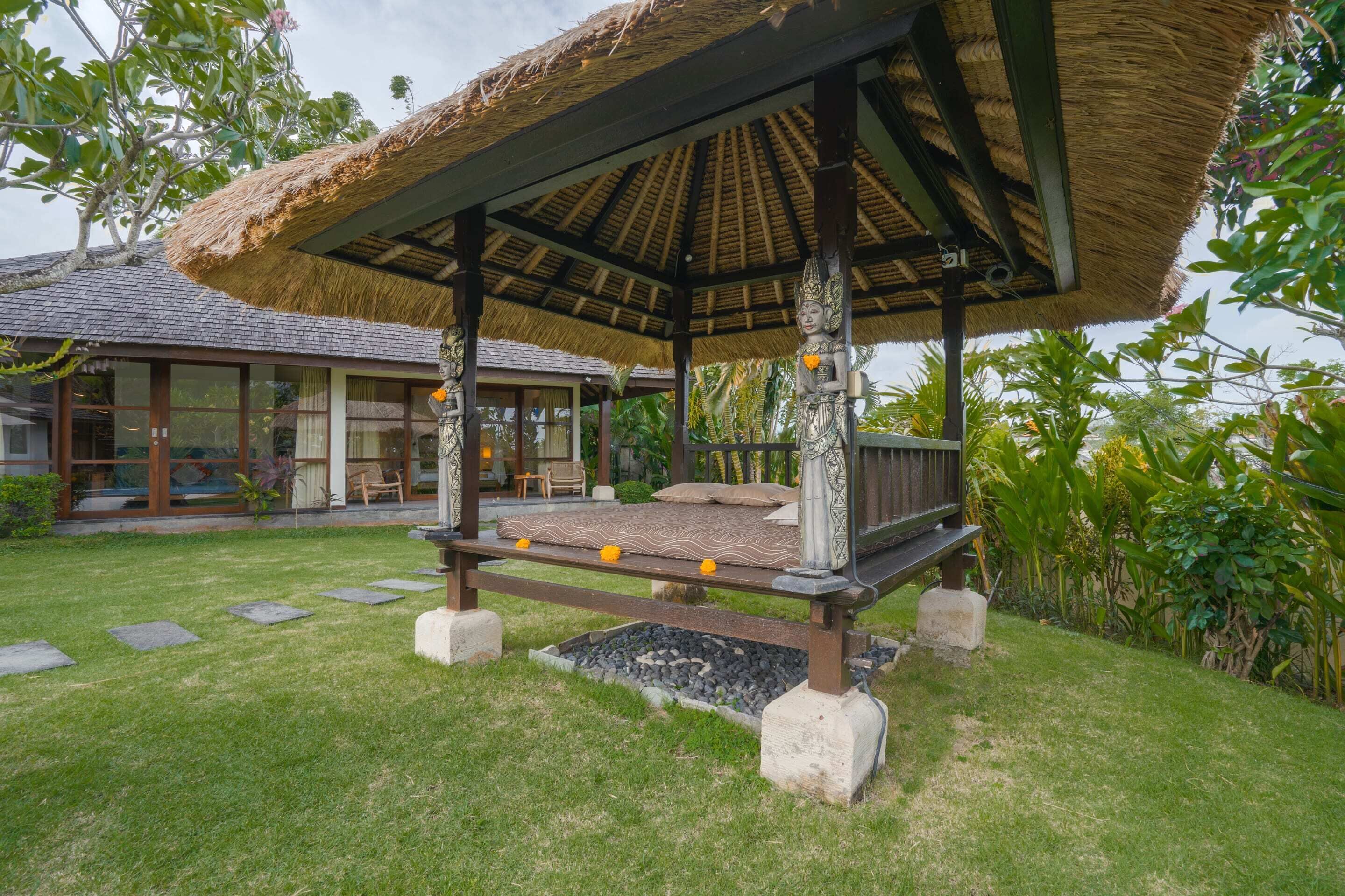 A thatched-roof gazebo with a bench, surrounded by a well-maintained lawn and tropical plants.