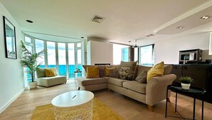 A modern living room with a sectional sofa, a round coffee table, and a view of the ocean through large windows. - Stunning Panoramic Sea View Beach Location 2 bed (bournemouth)