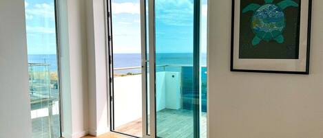 A room with a large sliding glass door leading to a balcony with a view of the ocean.