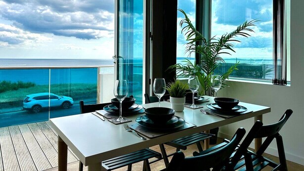 A dining area with a table set for two, large windows with a view of the sea and a car, and a potted plant on the windowsill. - Stunning Panoramic Sea View Beach Location 2 bed (bournemouth)